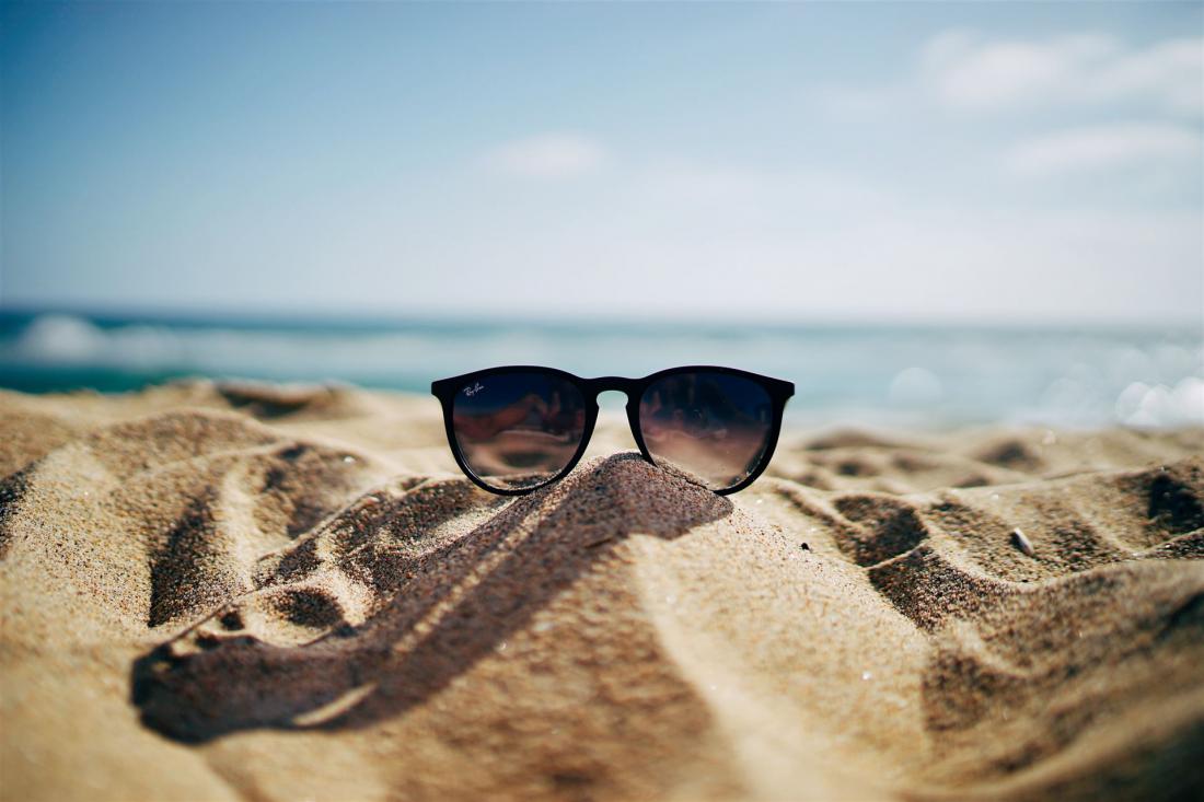 Eine Sonnenbrille liegt auf einem Sandhügel am Strand. Im Hintergrund sind das Meer und ein blauer Himmel zu sehen.