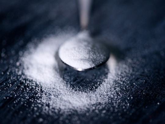 Ein Löffel mit weißem Pulver, vermutlich Zucker oder Salz, liegt auf einer dunklen Oberfläche. Das Pulver ist teilweise auf dem Löffel und teilweise auf der Oberfläche verstreut.